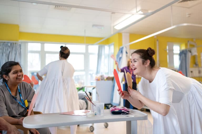 A woman in a white costume holds red props and smiles, bringing the joy of festivals as she interacts with a seated healthcare worker who is laughing in a bright hospital ward. Another woman in white observes the lively happenings in the background.